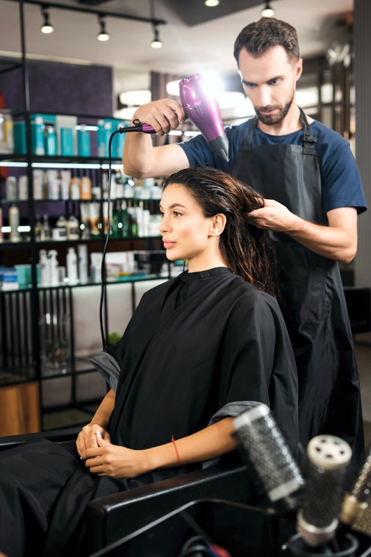 young hairdresser drying hair of woman near combs on blurred foreground